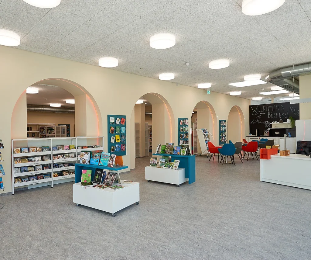 Heller Bibliotheks- und Aufenthaltsbereich einer Kita mit niedrigen Bücherregalen, runden Durchgängen, Sitzmöbeln und gleichmäßig beleuchteter Decke.