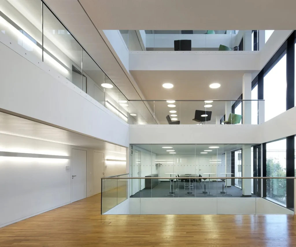 Moderner Innenraum eines Verwaltungsgebäudes mit mehreren Ebenen, Glasgeländern, offenen Galerien und gleichmäßig angeordneten Deckenleuchten