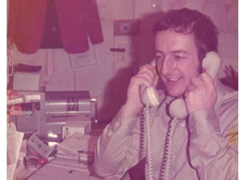 Ein Mann sitzt an einem Schreibtisch und telefoniert mit einem Festnetztelefon in einem Büro aus früherer Zeit